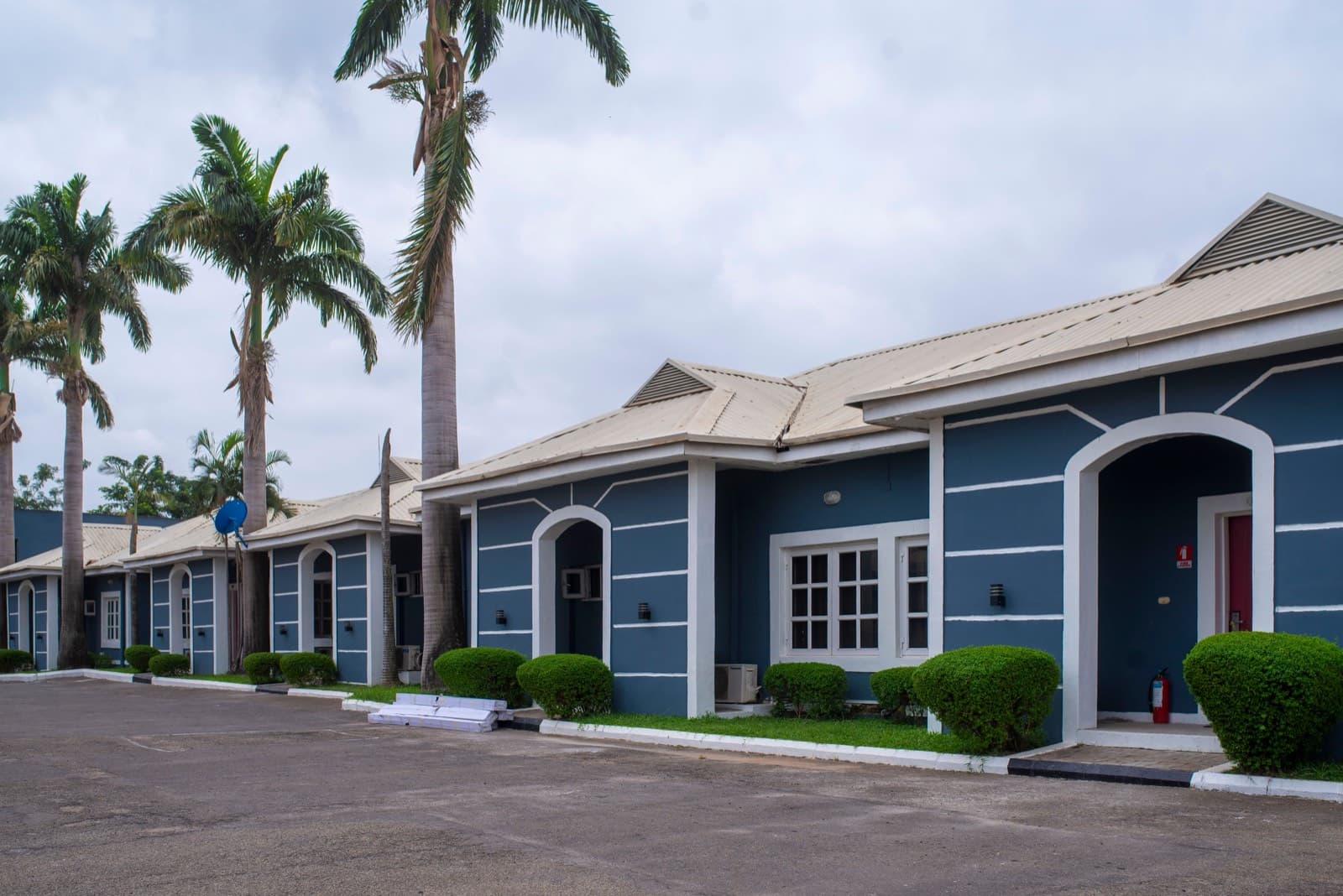 A row of blue and white single-storey residential units behind clipped hedges, with palm trees and wall-mounted air conditioning units